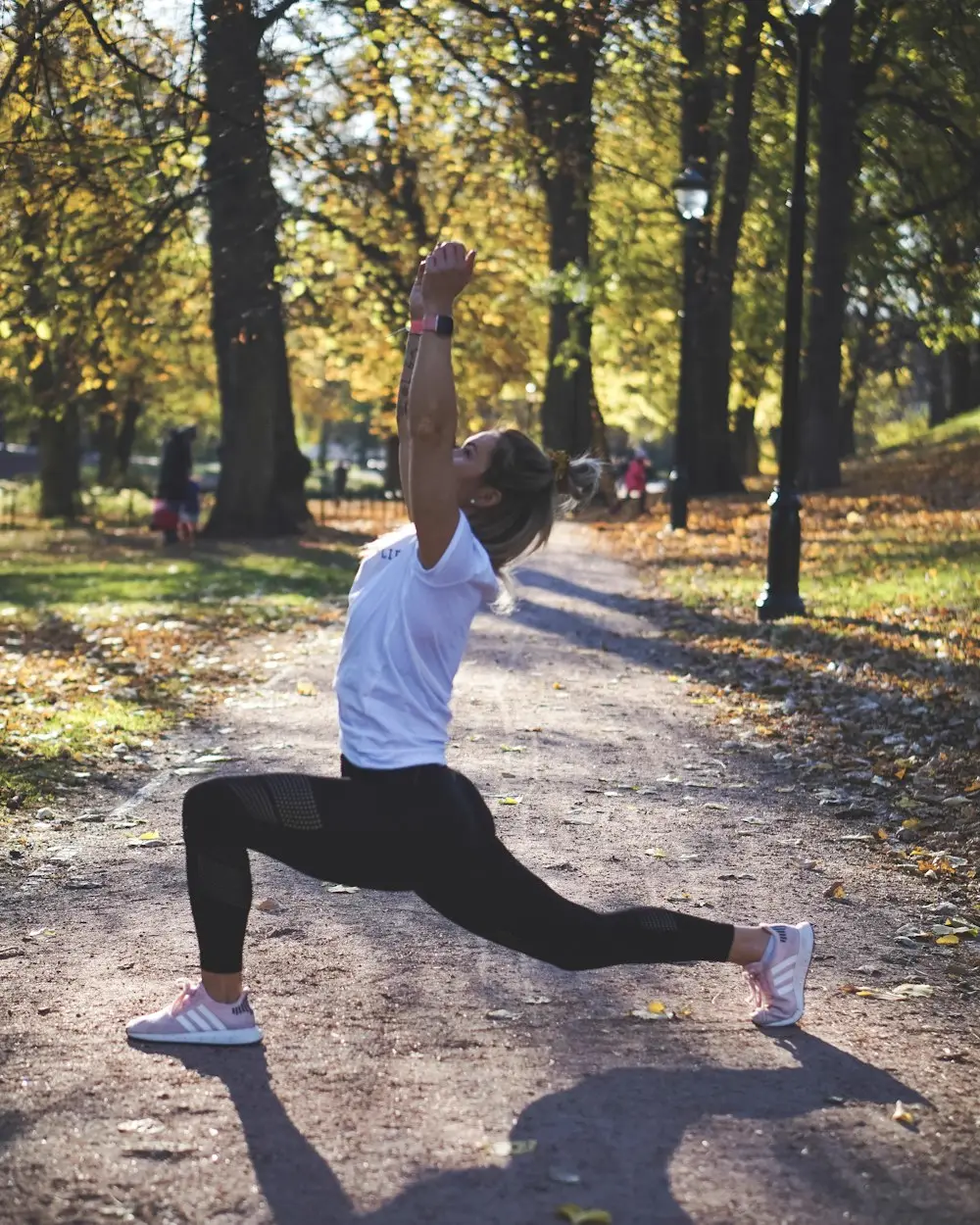 Une femme fait une fente bras levés sur une allée de parc couverte de feuilles mortes